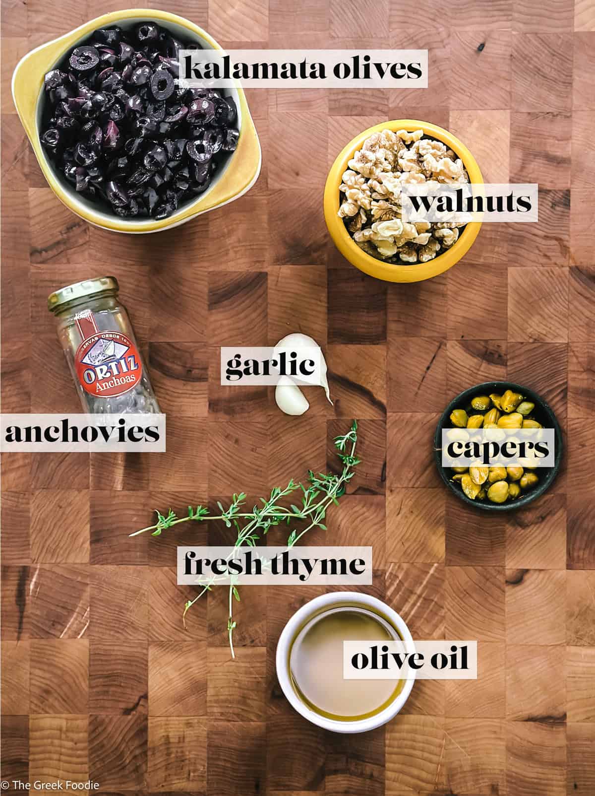 Flat lay of kalamata olive tapenade ingredients on a wooden chopping board, labeled: black kalamata olives, walnuts, anchovies, garlic, capers, fresh thyme, and olive oil