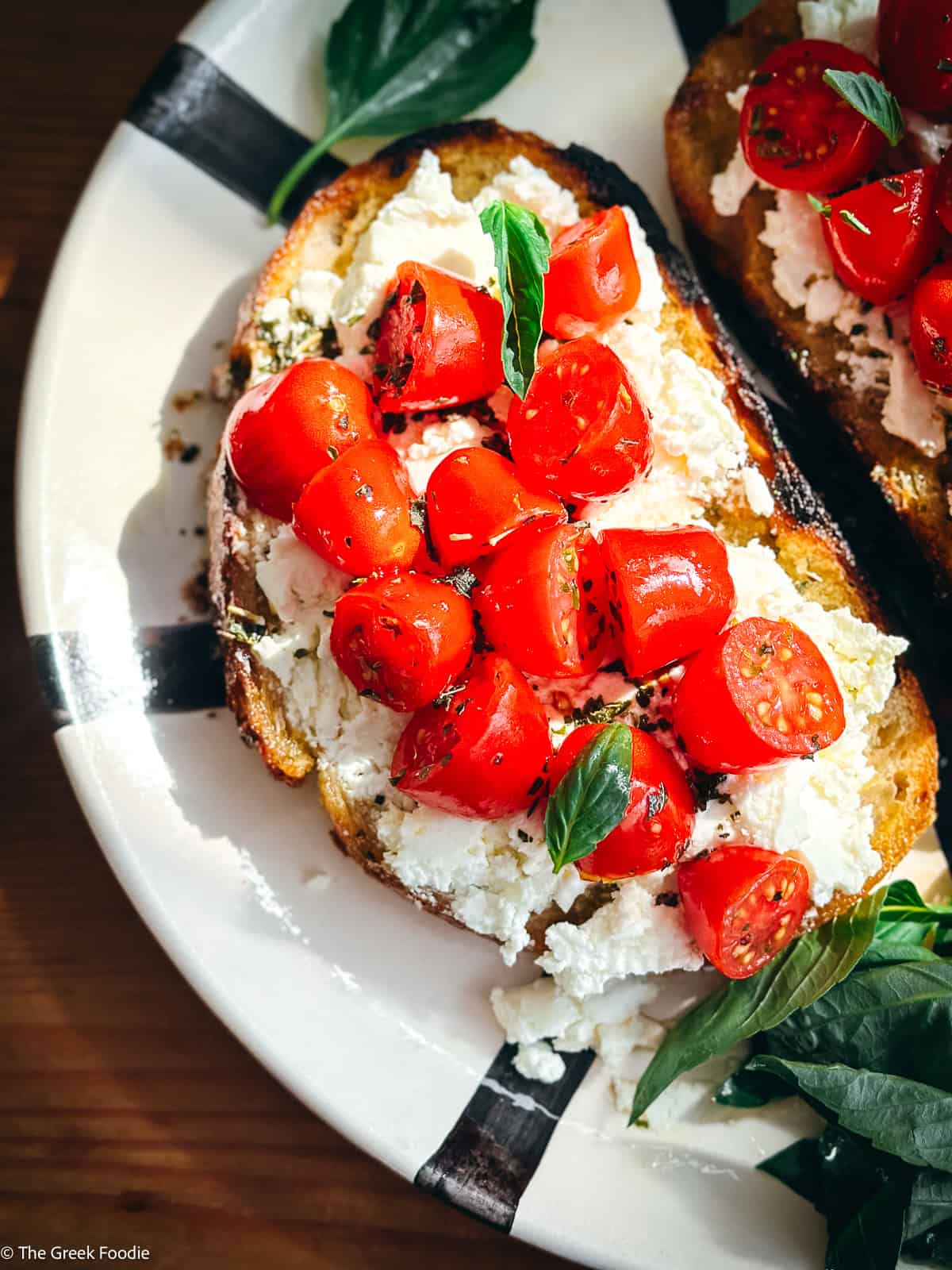Overhead view of tomato toast on a white plate with cherry tomatoes and fresh basil on a black and white surface