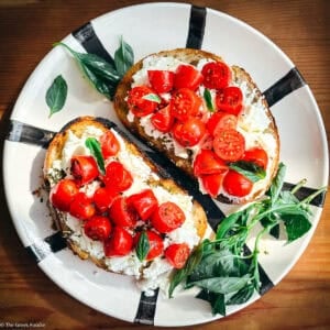 Tomato toast with mizithra and cherry tomatoes on a plate surrounded by fresh basil leaves