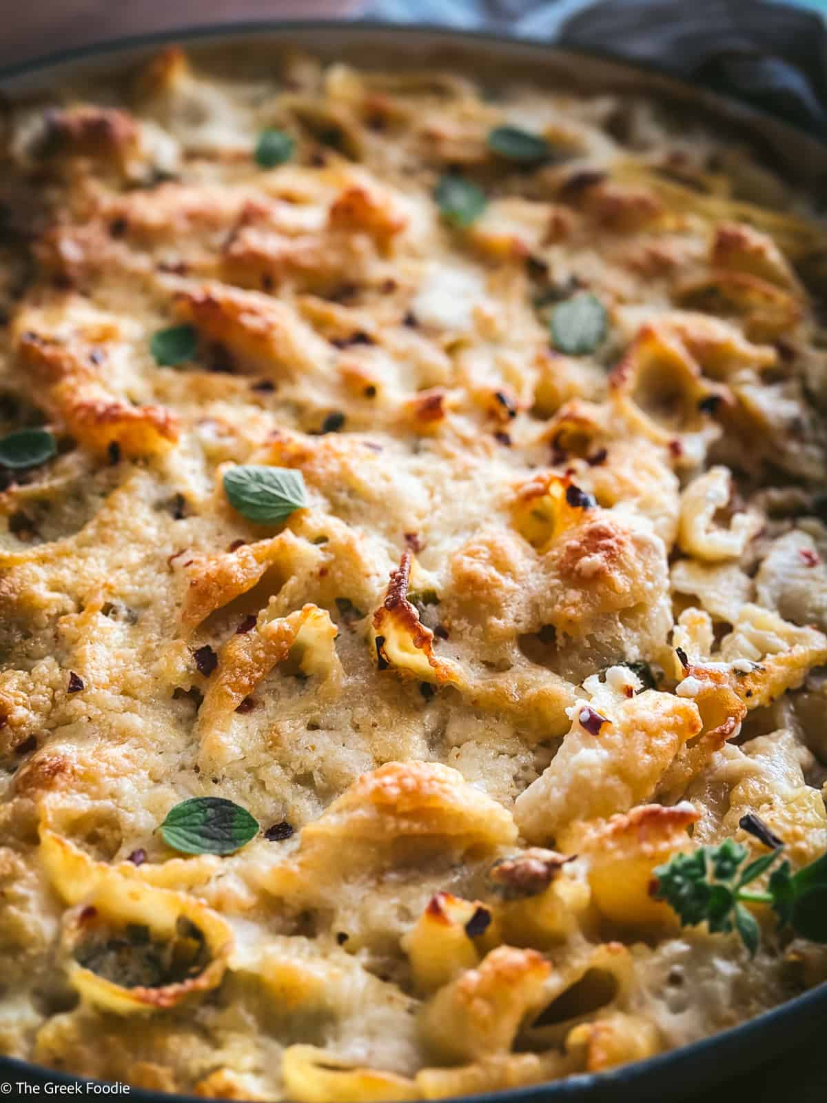 A close-up of baked Greek mac and cheese showing the golden, bubbling graviera crust with shell pasta, fresh oregano, and chilli flakes visible throughout.