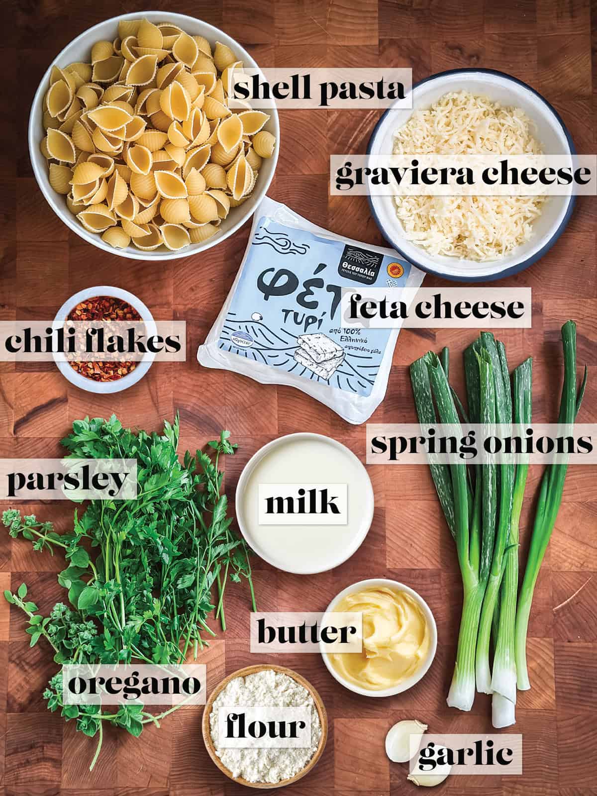 All the ingredients for Greek mac and cheese laid out on a wooden board — shell pasta, grated graviera, a packet of Greek feta, chilli flakes, spring onions, fresh parsley, fresh oregano, milk, butter, flour, and garlic.