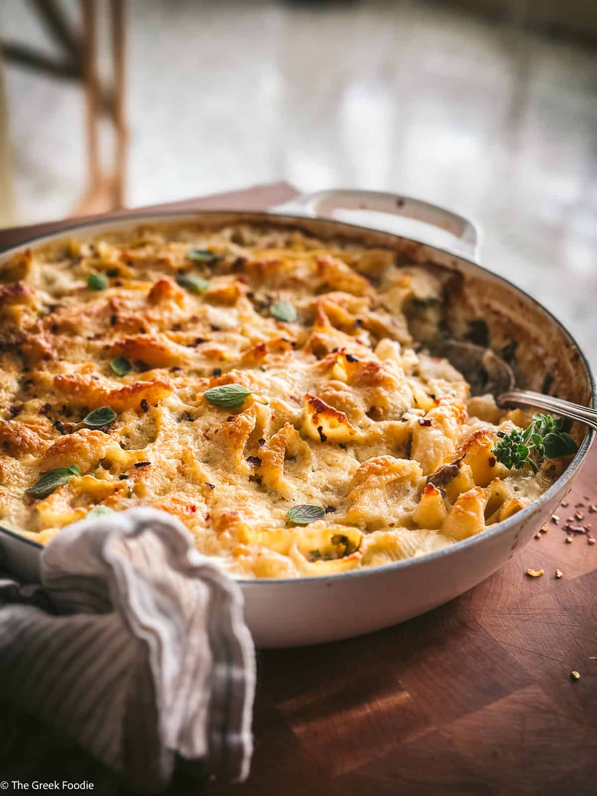 A wide pan of Greek mac and cheese with feta and graviera on a wooden table, the golden cheese crust bubbling and topped with fresh oregano, a serving spoon resting in the pan.