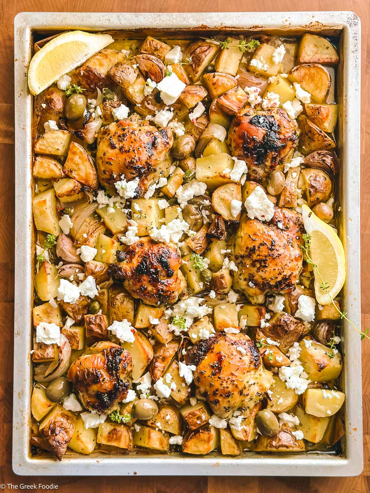 Greek sheet pan chicken and potatoes with feta on a wooden butcher block, garnished with lemon