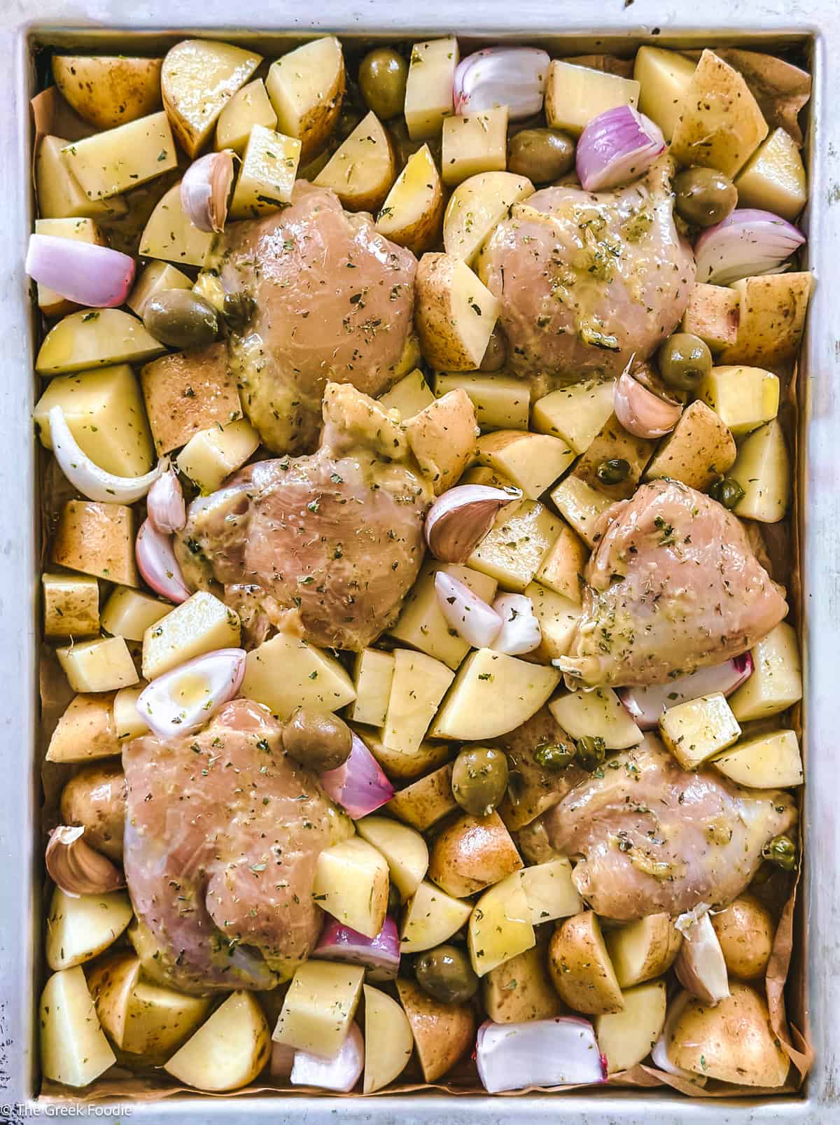 Greek sheet pan chicken assembled and ready for the oven, with potatoes, onions, and capers