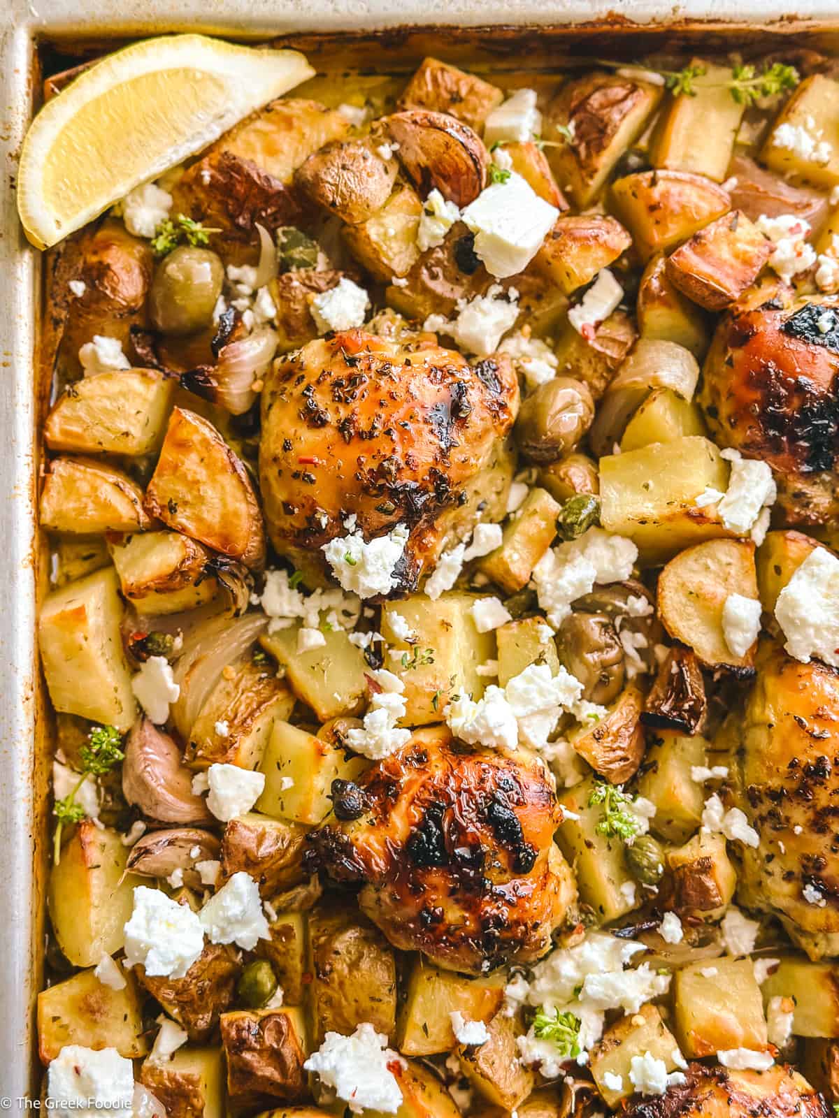 Greek sheet pan chicken thighs roasted with potatoes, crumbled feta, and a wedge of lemon.