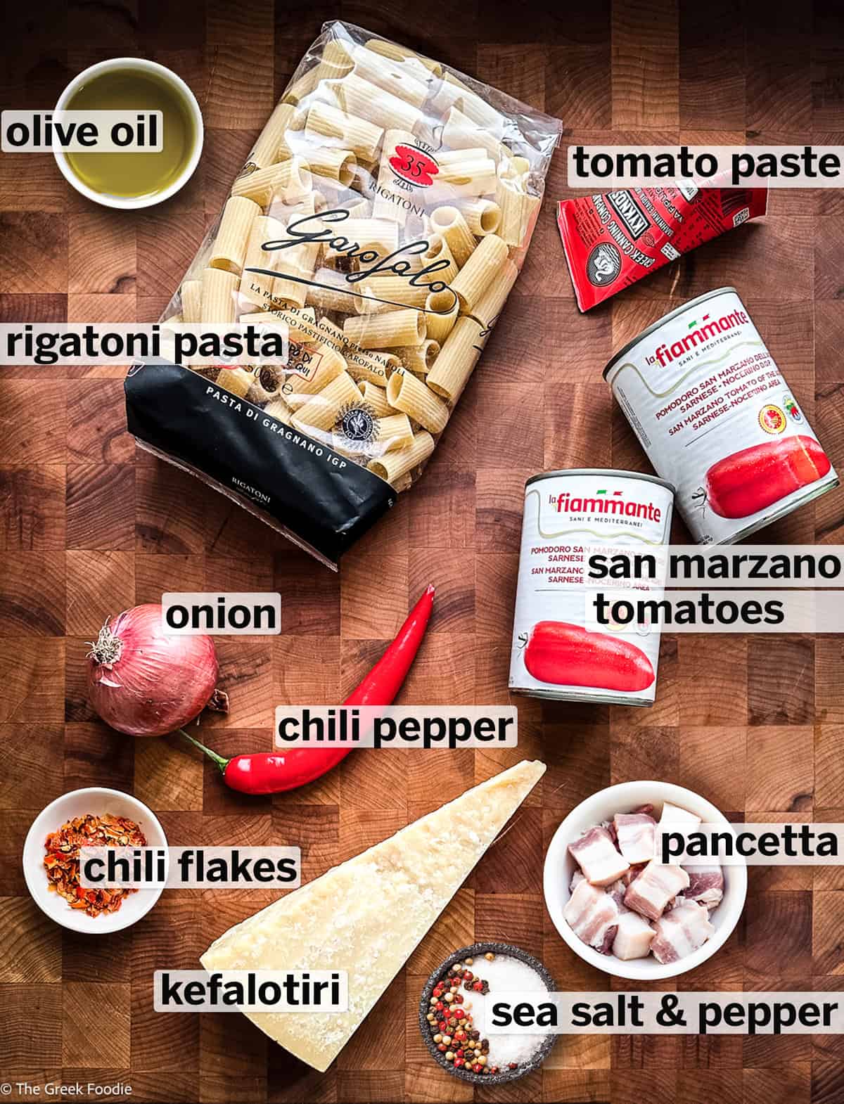 Labeled flat lay of ingredients for spicy tomato pasta: rigatoni, San Marzano tomatoes, tomato paste, pancetta, kefalotiri, onion, chili pepper, chili flakes, olive oil, and seasoning
