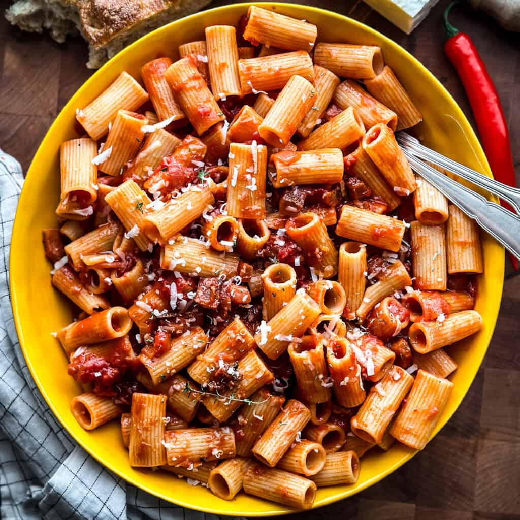 Overhead shot of a yellow bowl of spicy tomato rigatoni with a fork, a fresh red chili, and crusty bread on the side