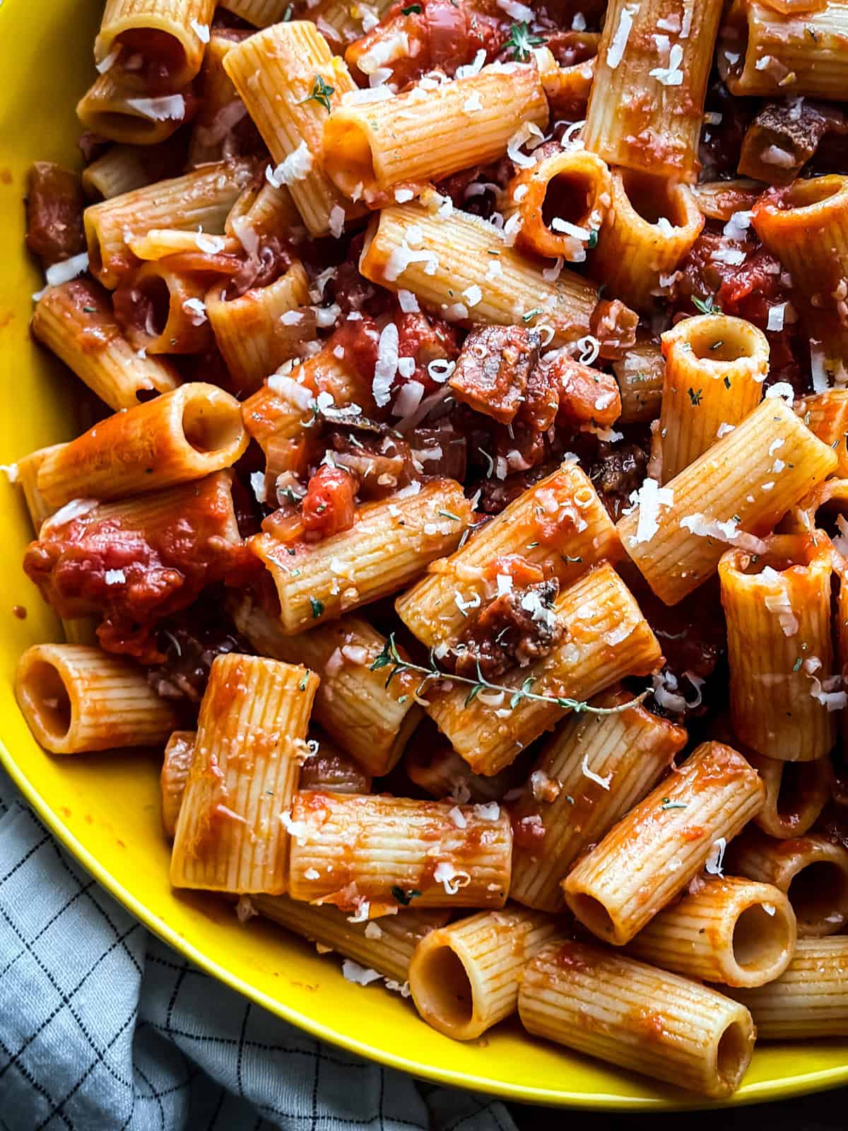 Close overhead view of rigatoni tossed in spicy tomato sauce with pancetta, kefalotiri, and thyme leaves in a yellow bowl