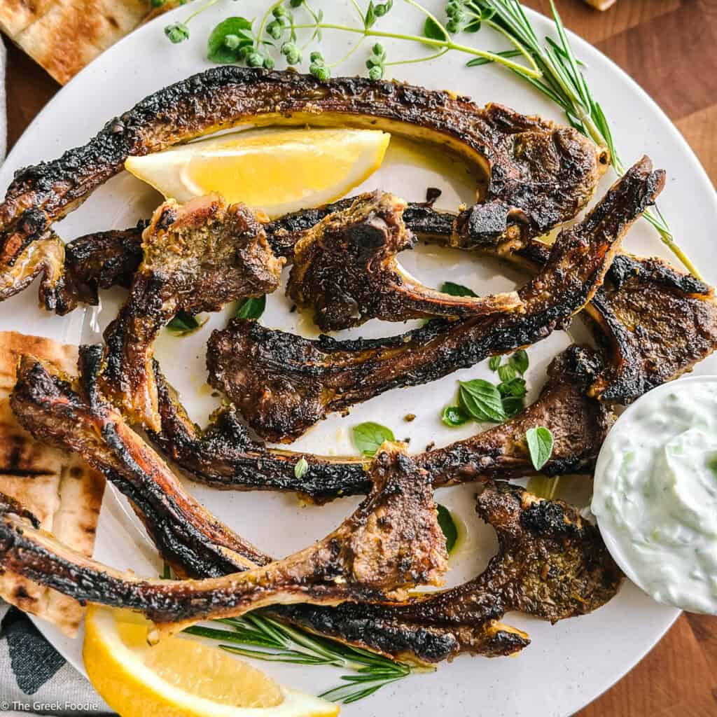 Overhead of grilled Greek lamb chops (paidakia) on a white plate with a lemon wedge, fresh oregano, rosemary, and a bowl of tzatziki