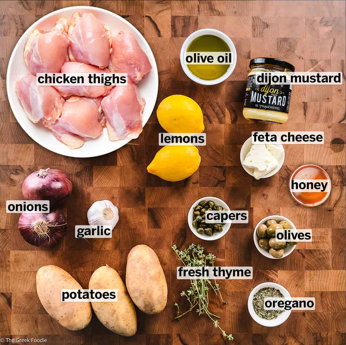 Gathered ingredients for Greek sheet pan chicken with labels on a butcher block shot from above.