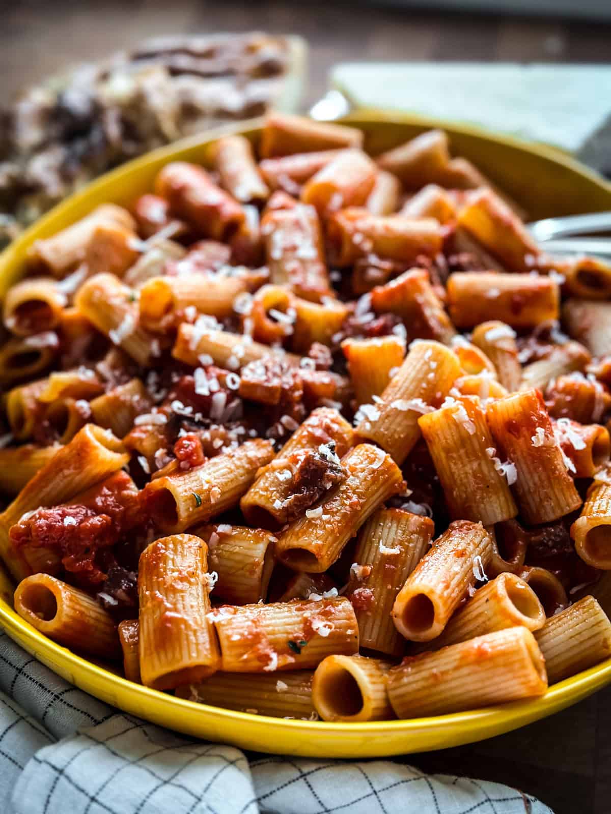 Yellow bowl of rigatoni coated in spicy tomato sauce with crisp pancetta and grated kefalotiri, served with a fork