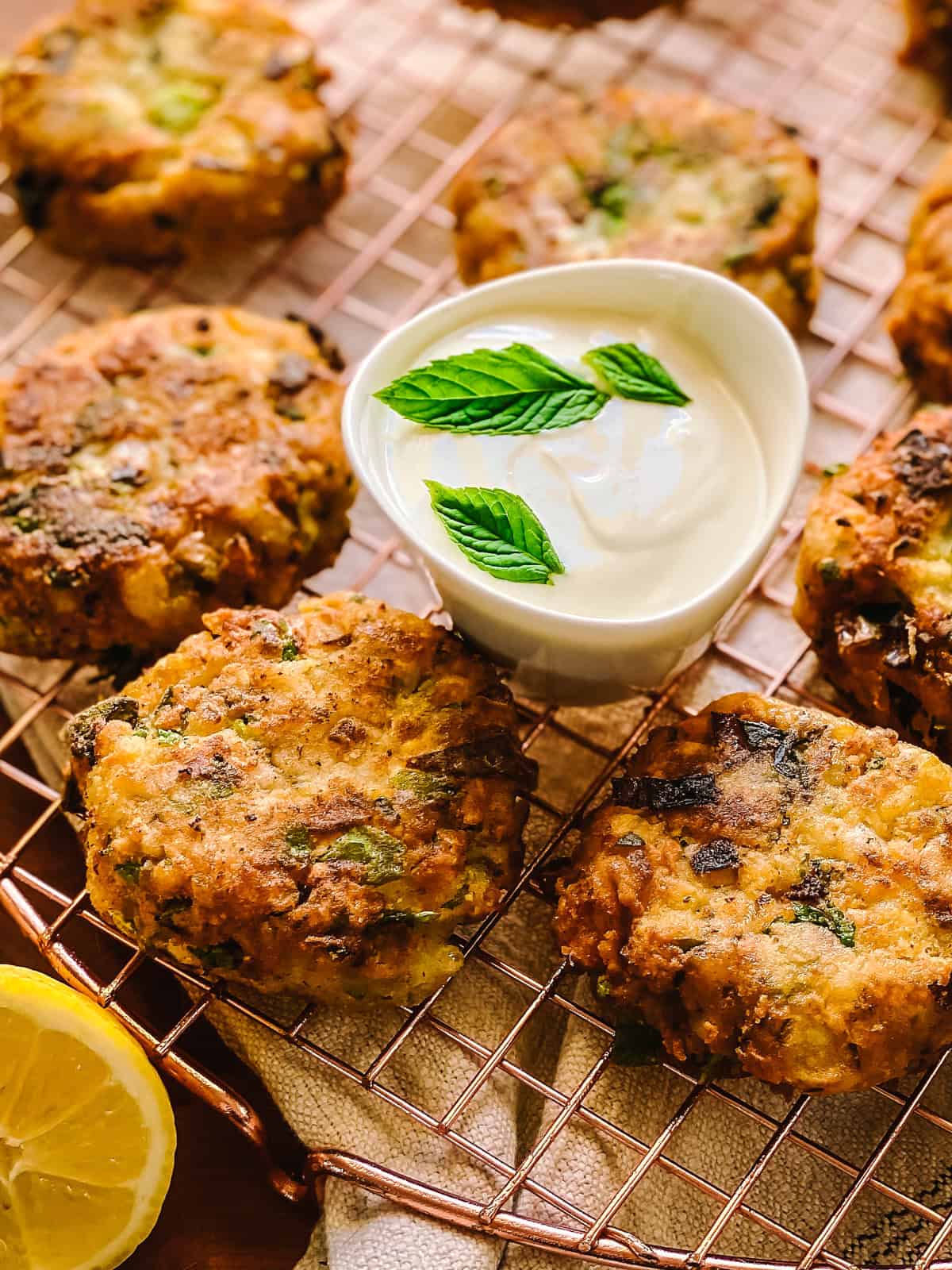 Greek chickpea fritters on a copper wire rack with a bowl of tahini lemon yogurt sauce and a fresh lemon half