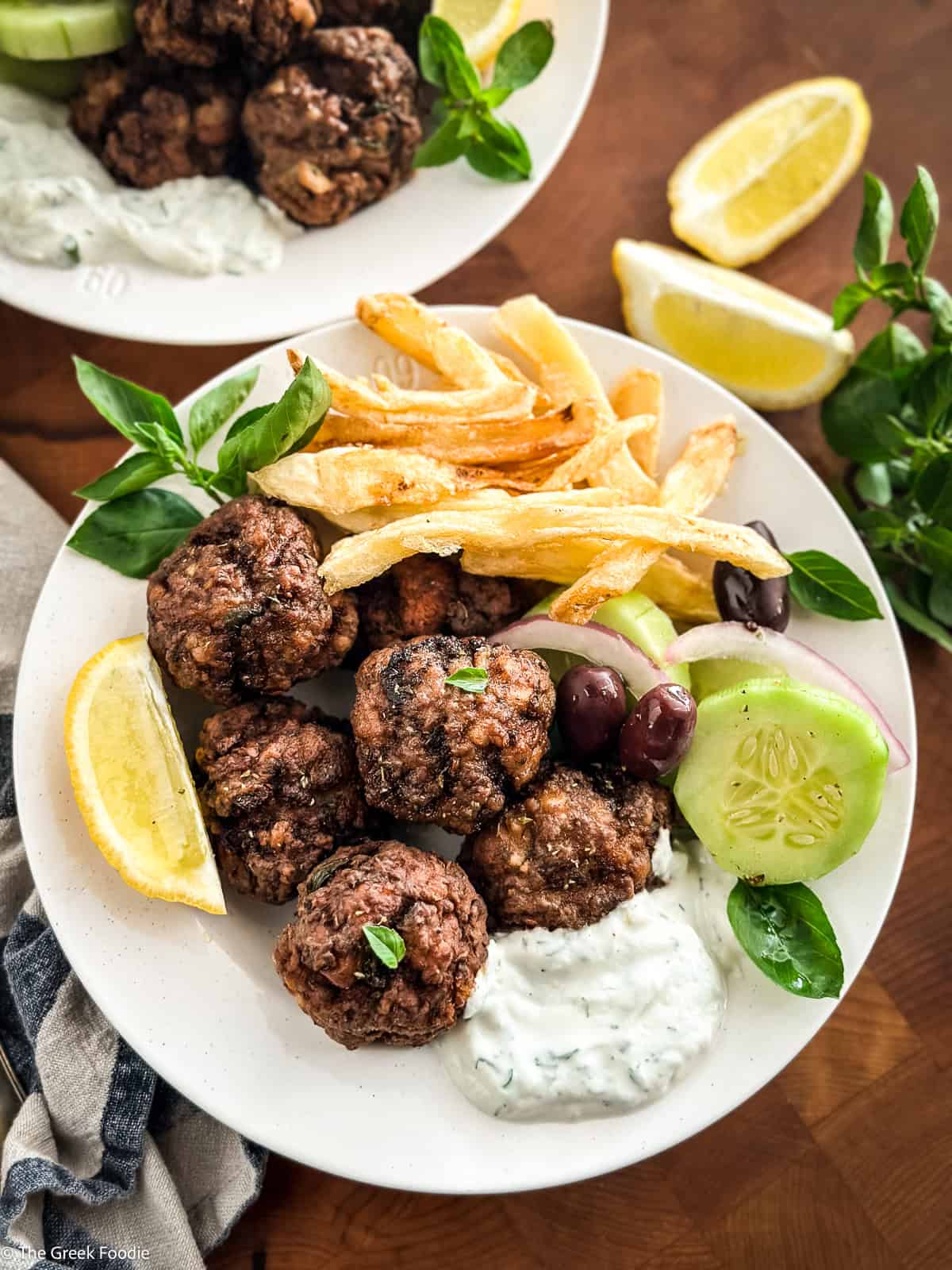 Taverna-style plate of Greek meatballs and fries with tzatziki, olives and lemon on a wooden table.