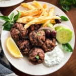 Greek meatballs served with tzatziki, crispy fries, cucumber, olives and a lemon wedge on a white plate.