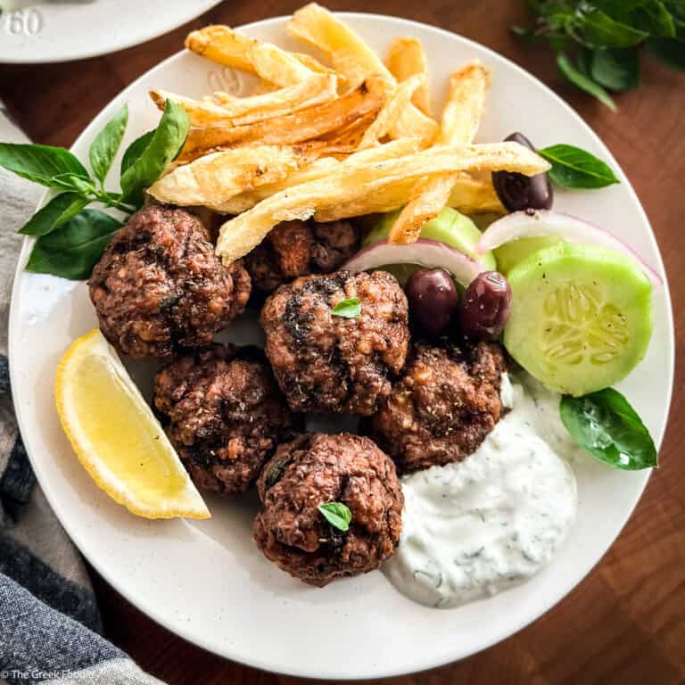Greek meatballs served with tzatziki, crispy fries, cucumber, olives and a lemon wedge on a white plate.