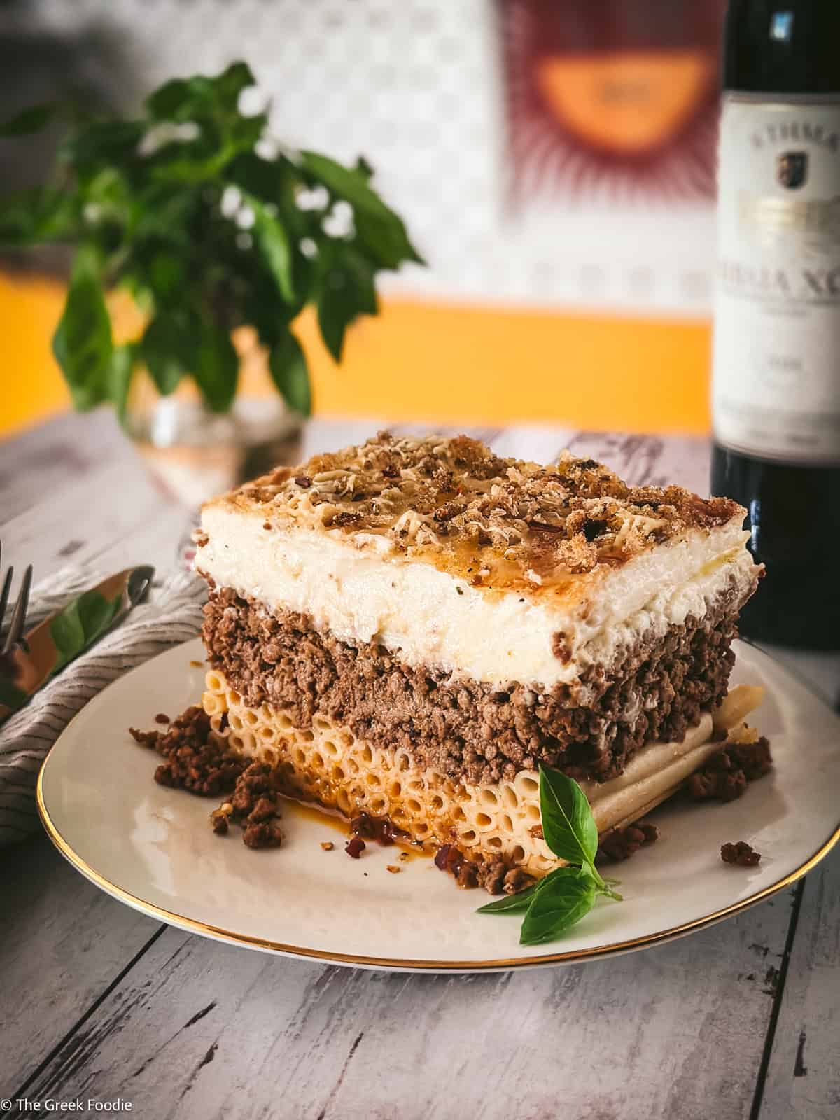 A tall slice of Greek pastitsio on a white plate with fresh herbs and a bottle of Greek wine behind