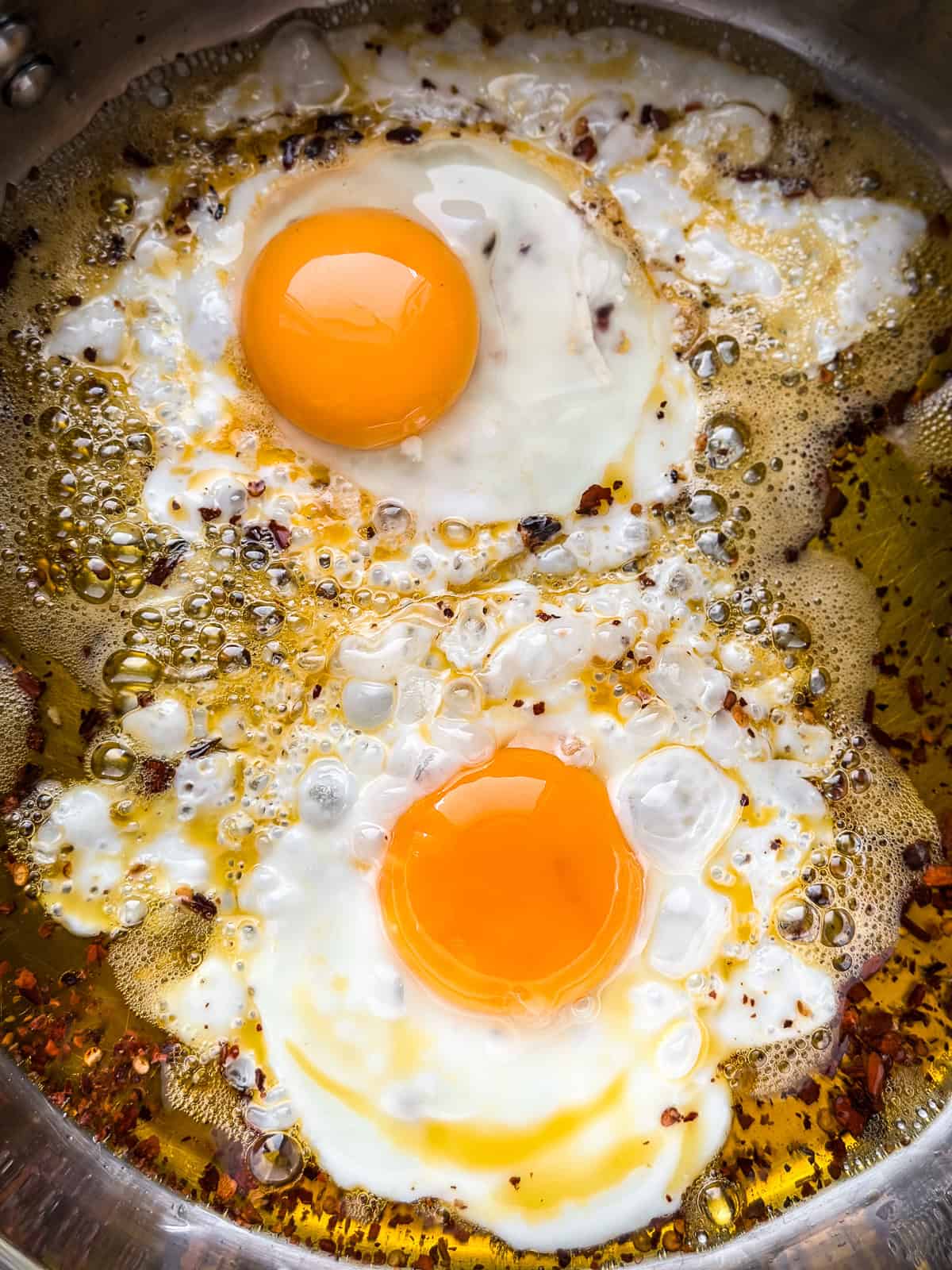 Two eggs frying in bubbling chili-infused olive oil with charred chili flakes, in a stainless steel pan