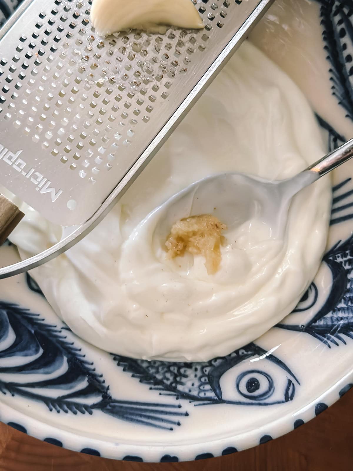 Grating fresh garlic into a spoonful of strained Greek yogurt in a blue and white patterned bowl