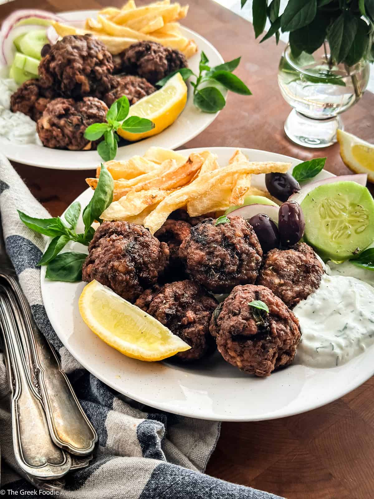 Two plates of Greek keftedes served meze-style with fries, olives, cucumber and tzatziki.