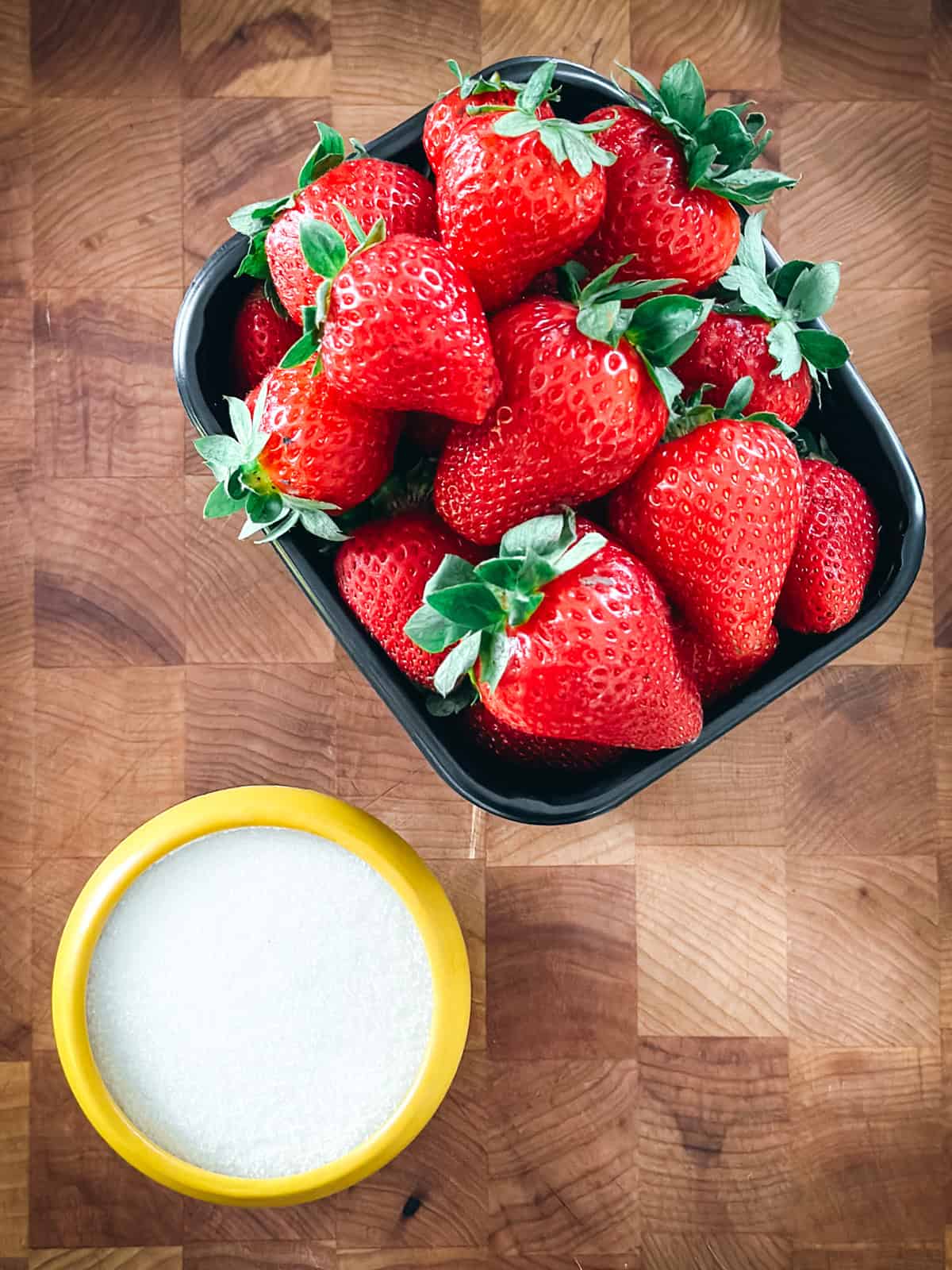 Fresh strawberries in a black bowl beside a bowl of sugar on a wooden board