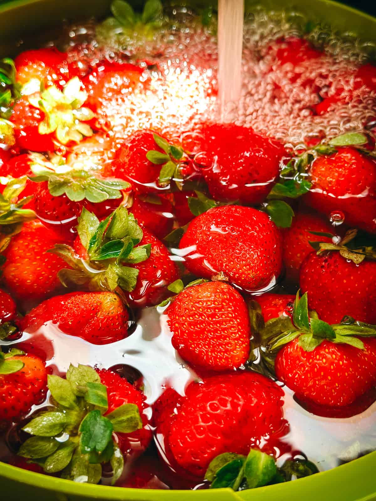 Fresh strawberries being rinsed in a green colander under running water