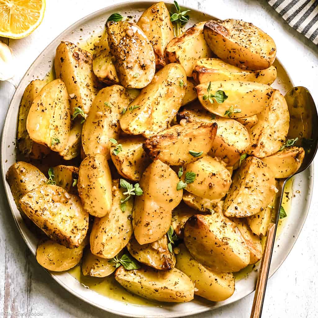 Crispy Greek lemon potatoes with garlic and oregano on a white platter, glossy with lemon pan sauce.