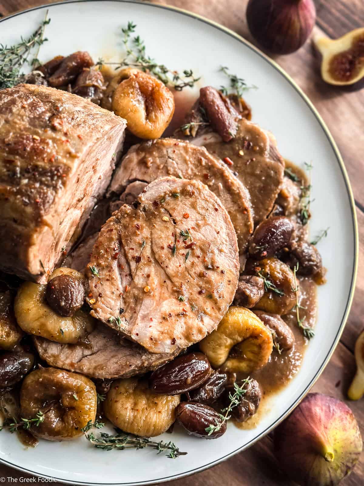 Sliced Greek pork pot roast on a white platter with plumped figs, dates and chestnuts in a glossy brown sauce