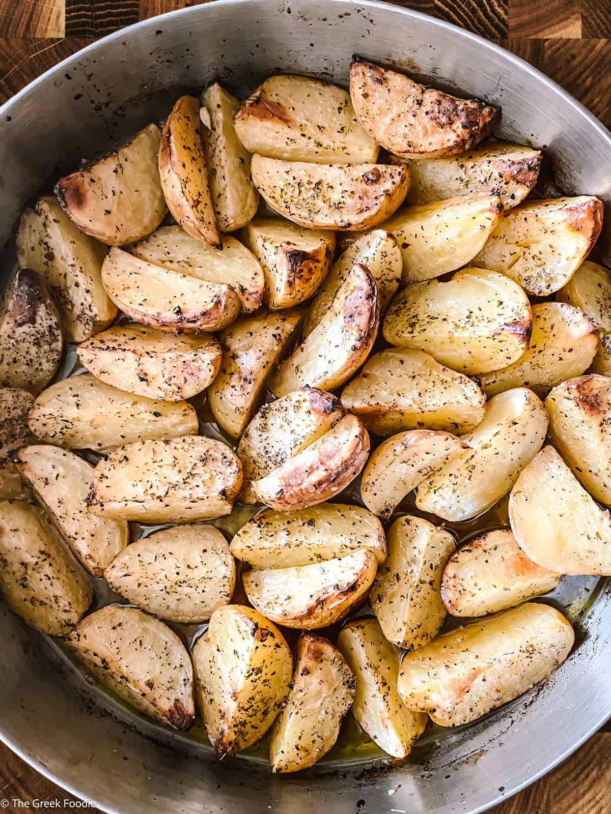 Greek potatoes roasting in a baking pan, edges turning golden brown in the lemon and olive oil sauce.