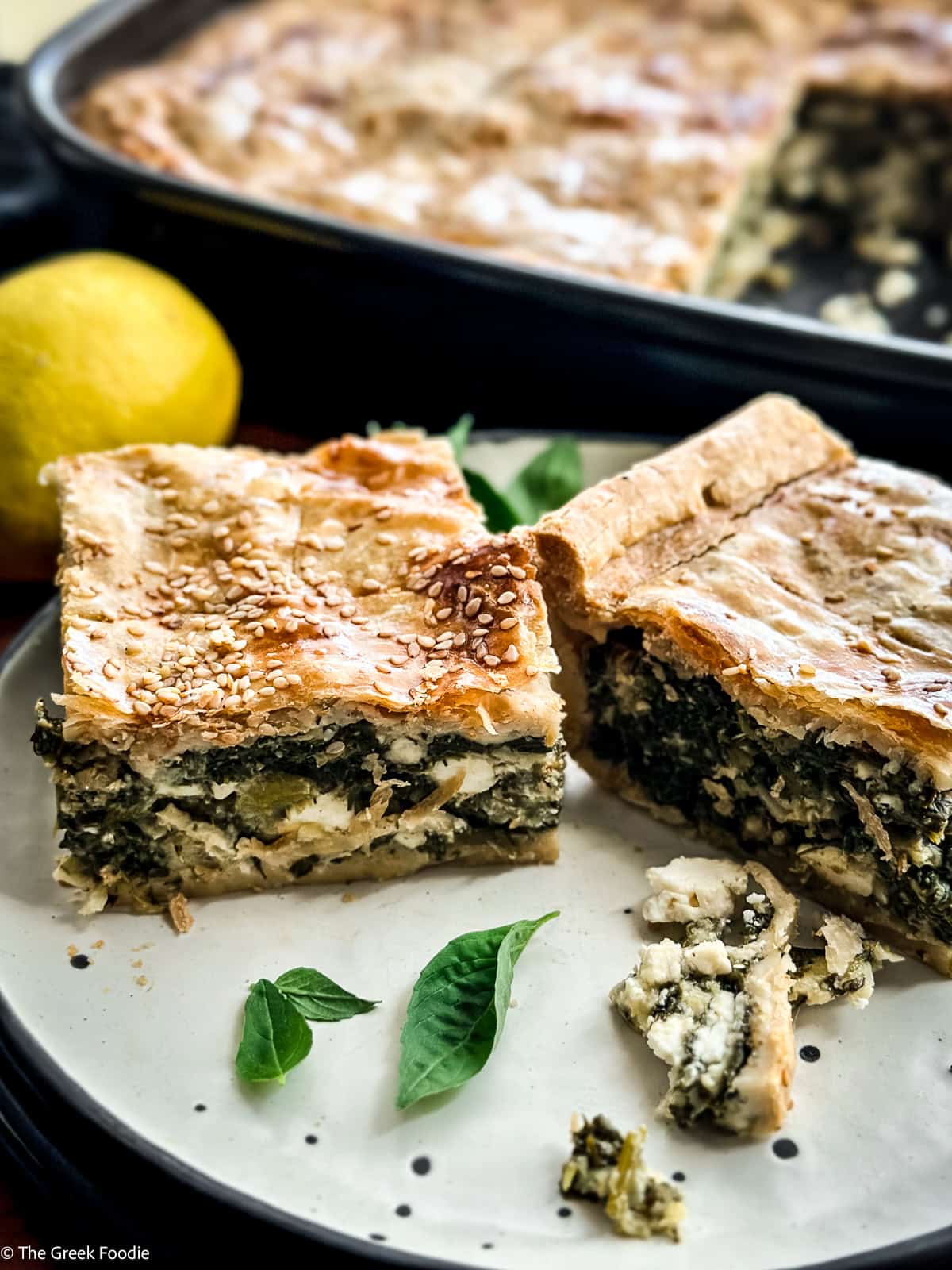 Squares of hortopita, Greek savory pie with greens and feta, on a plate with basil leaves and a lemon.