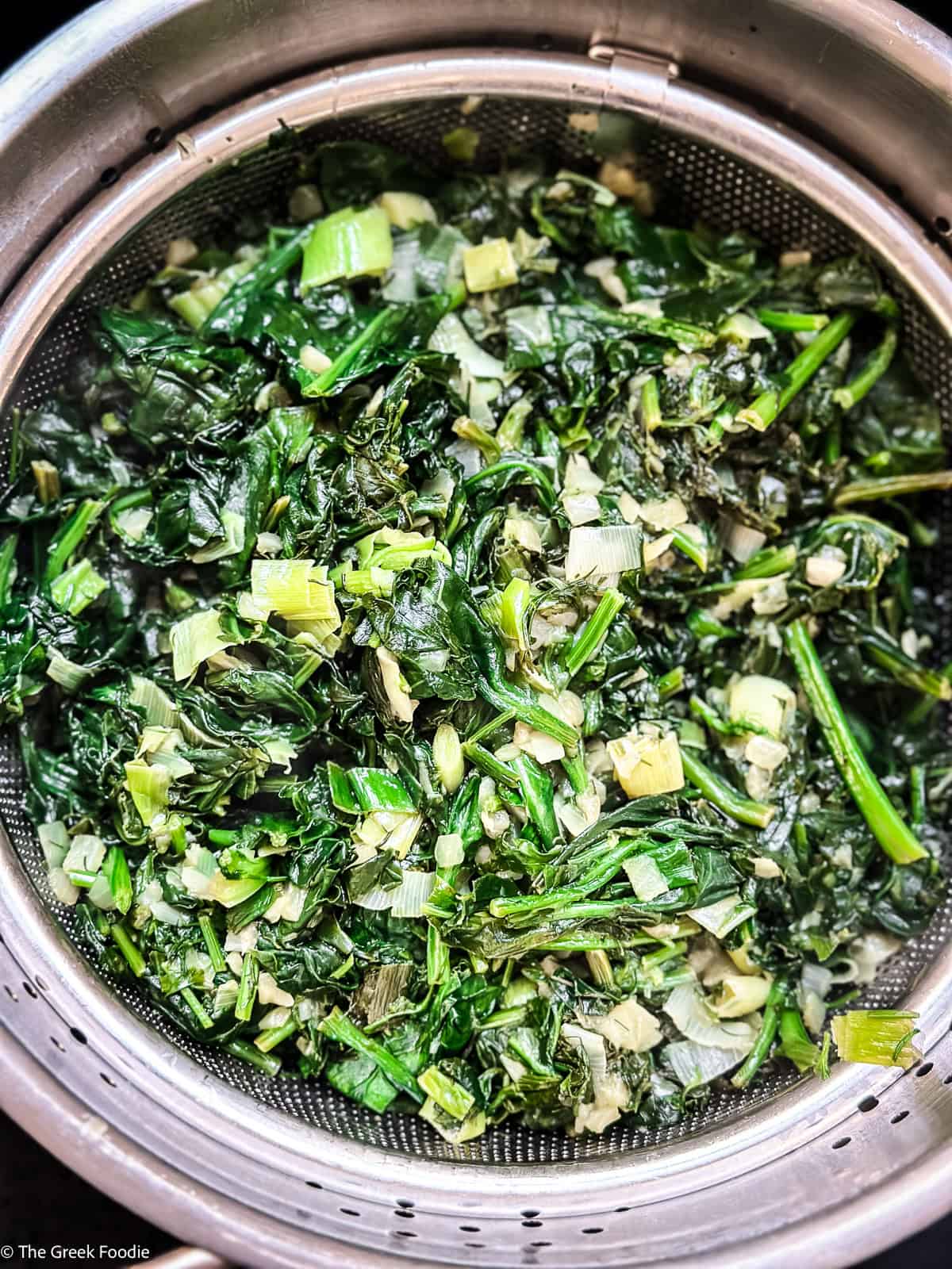 Cooked greens draining in a metal colander before being mixed into the hortopita filling.