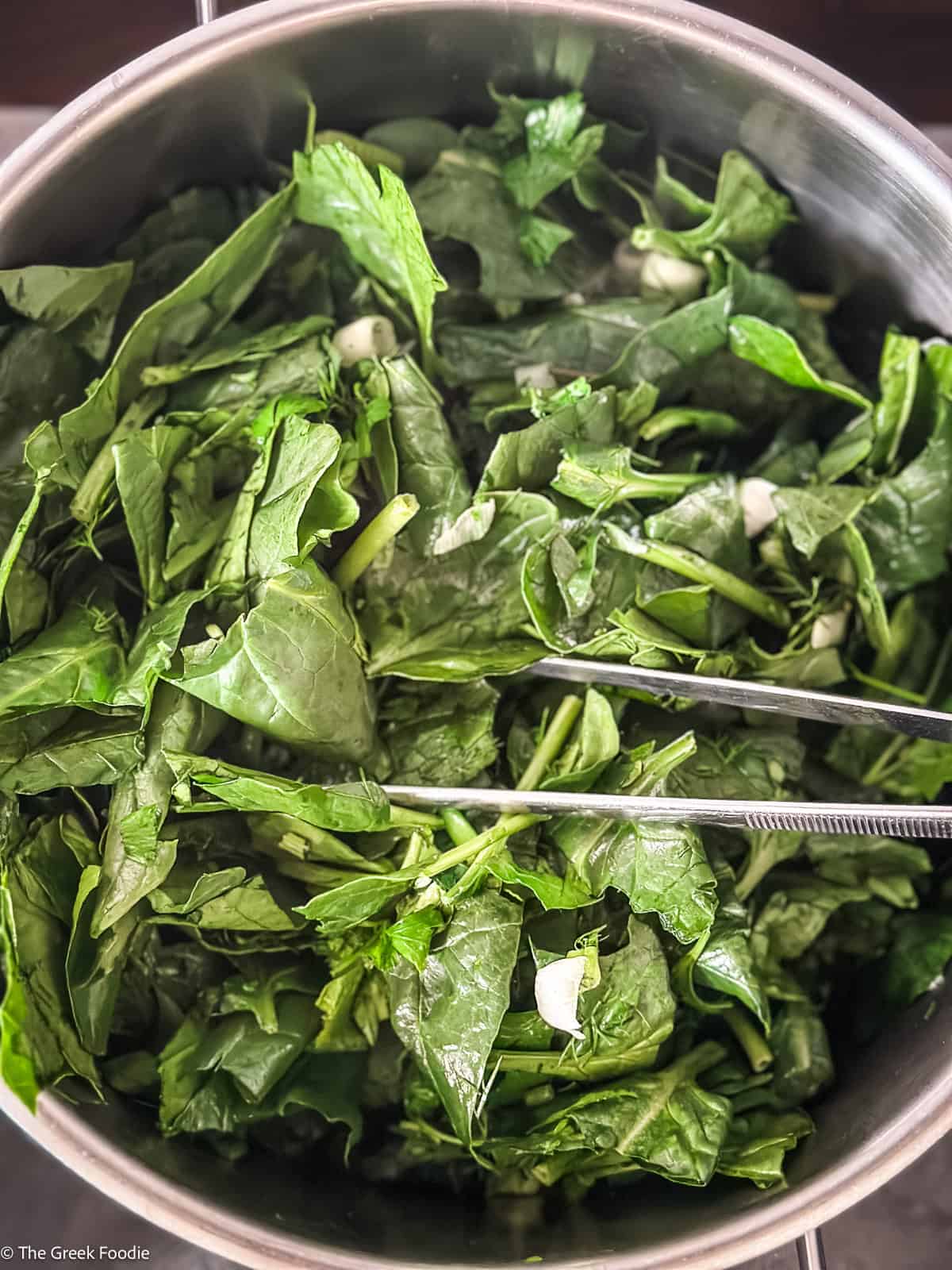 Chopped greens wilting in a pot with tongs, the first step in making hortopita.