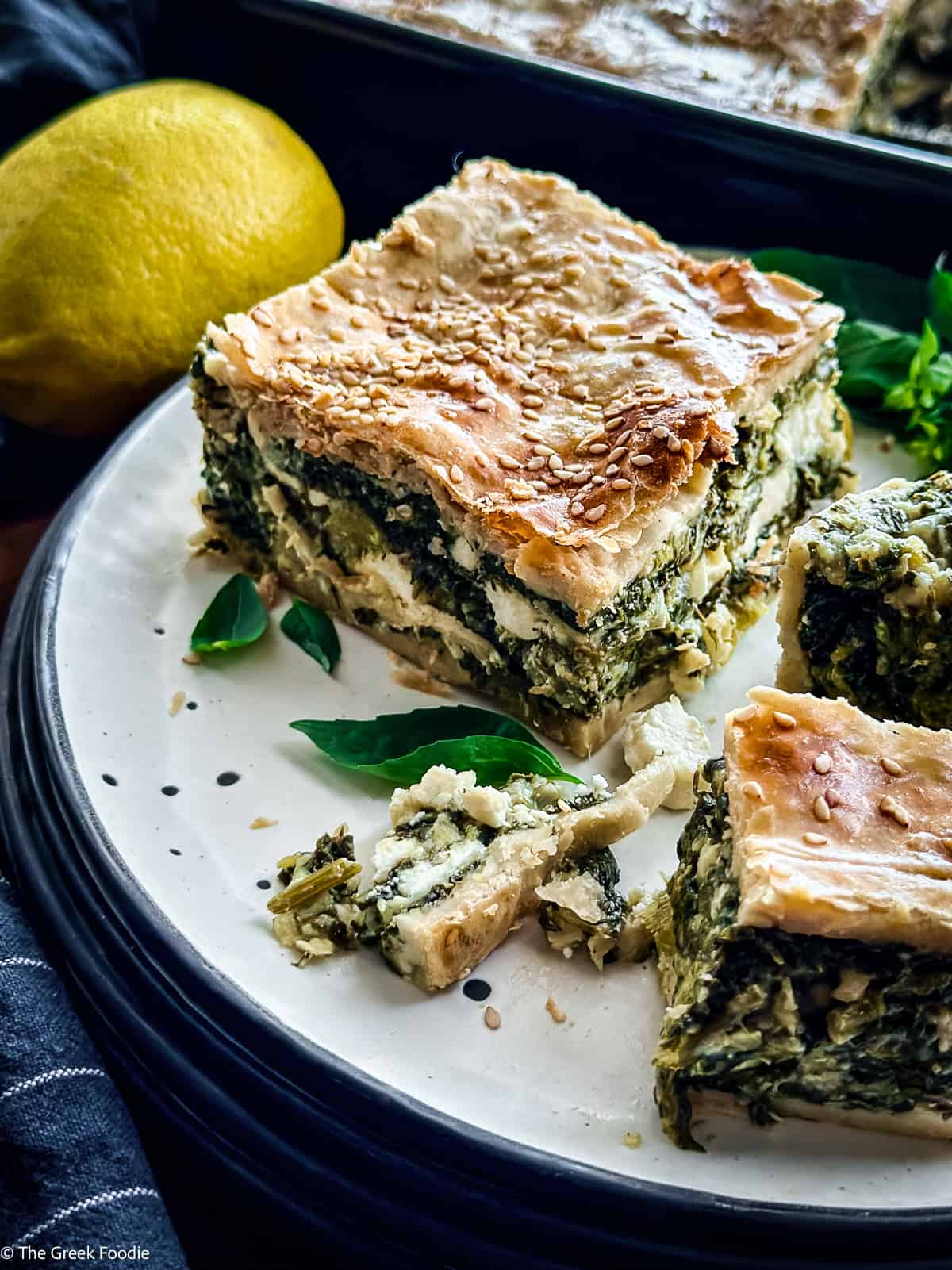 Two squares of hortopita on a white plate with fresh basil and a lemon, the baking pan behind.