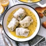 Four lahanodolmades in a white bowl of golden avgolemono sauce, with lemon, and crusty bread