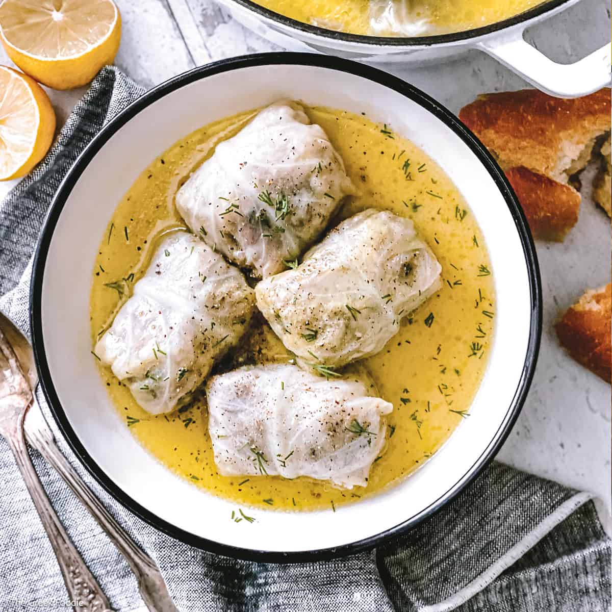 Four lahanodolmades in a white bowl of golden avgolemono sauce, with lemon, and crusty bread
