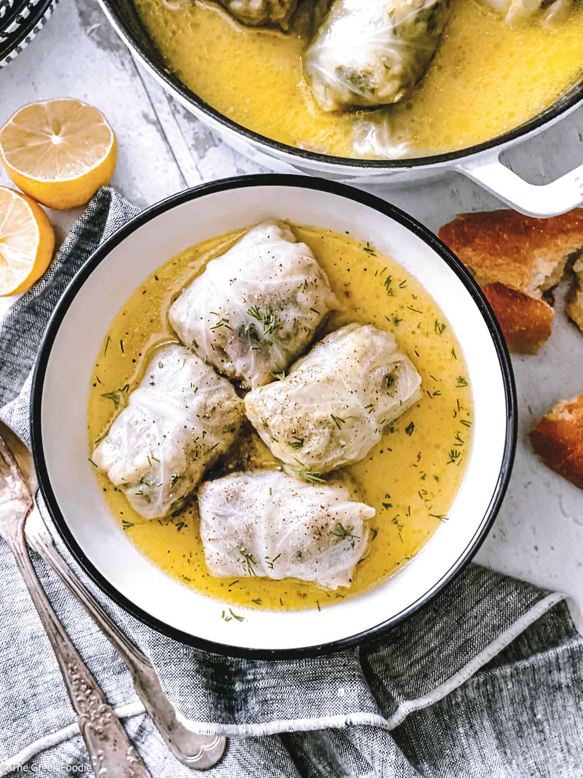 Four lahanodolmades in a white bowl of golden avgolemono sauce, with lemon, crusty bread and a linen napkin