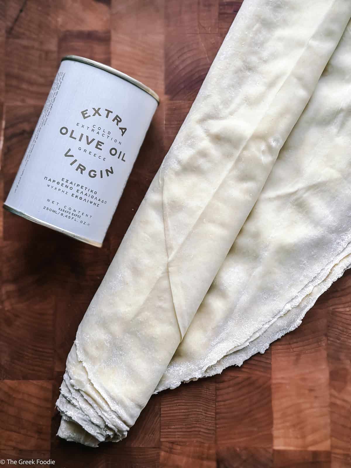 A roll of country-style phyllo dough beside a can of Greek extra virgin olive oil on a wooden board.