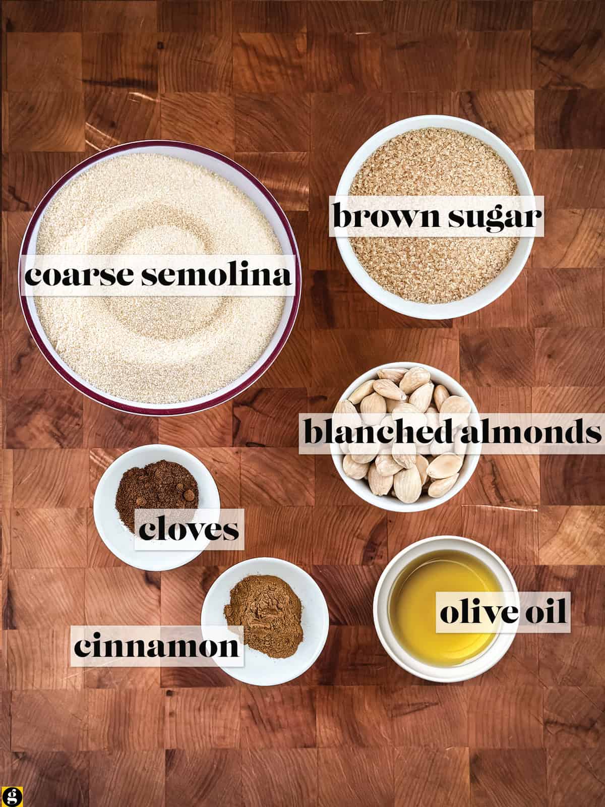 Ingredients for Greek halva in bowls on a wooden board: coarse semolina, brown sugar, blanched almonds, olive oil, ground cinnamon, and ground cloves