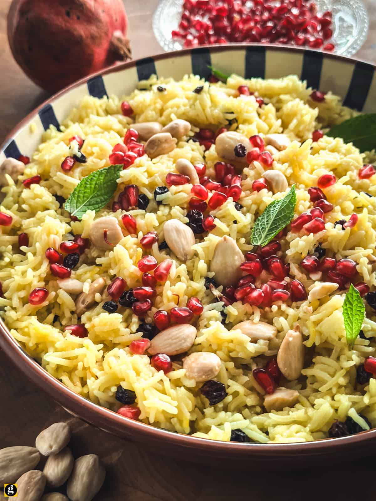 Greek saffron rice pilaf in a striped bowl, topped with pomegranate seeds, blanched almonds, currants and fresh mint.
