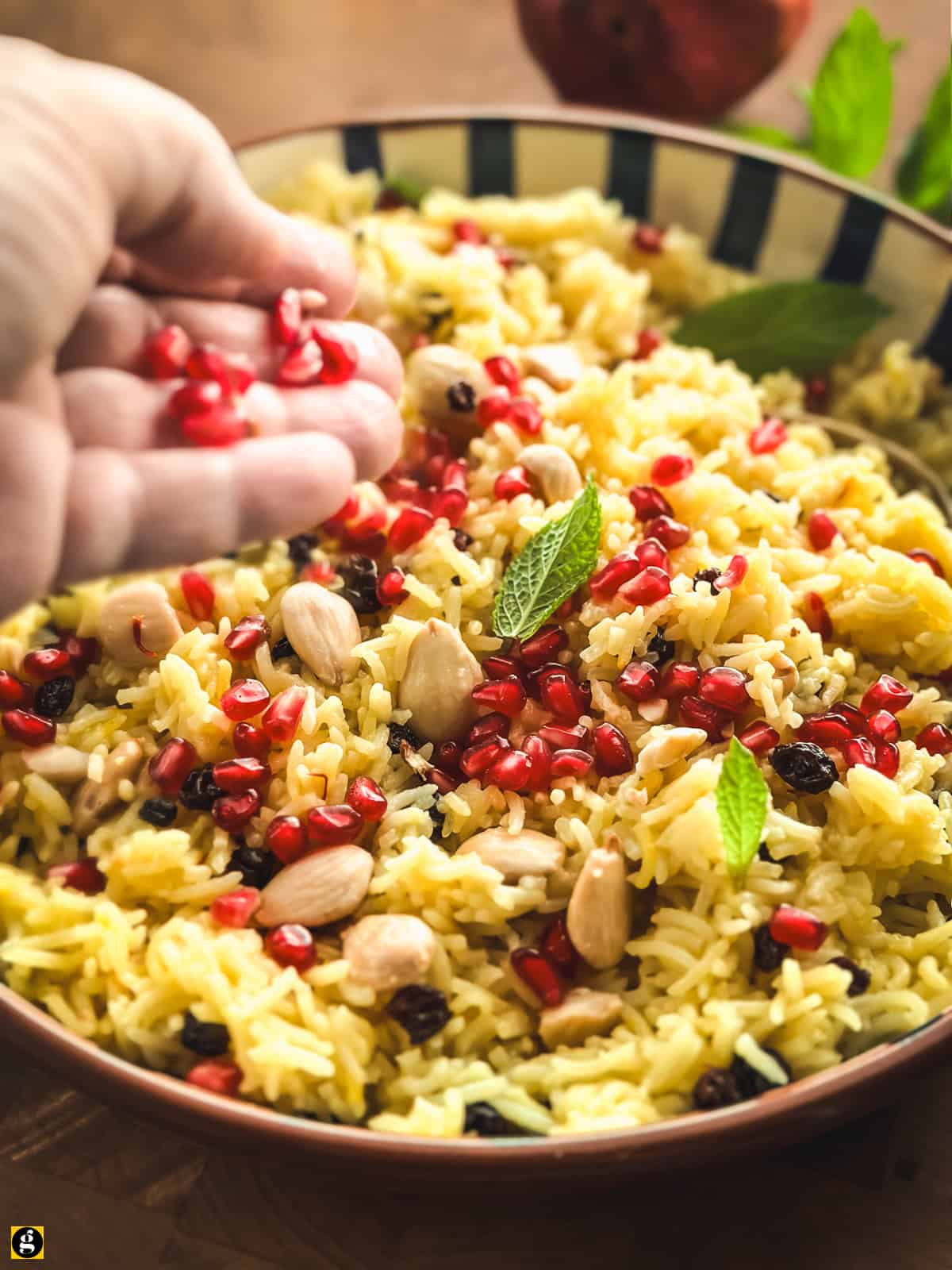 A hand scattering pomegranate seeds over a bowl of Greek saffron rice pilaf.