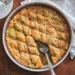 Overhead view of Greek pistachio baklava in a round metal pan with a serving spoon, glass plates beside the baking pan