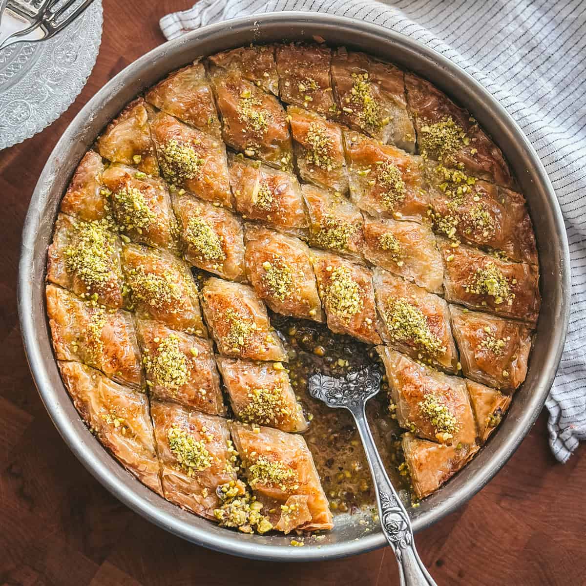Overhead view of Greek pistachio baklava in a round metal pan with a serving spoon, glass plates beside the baking pan