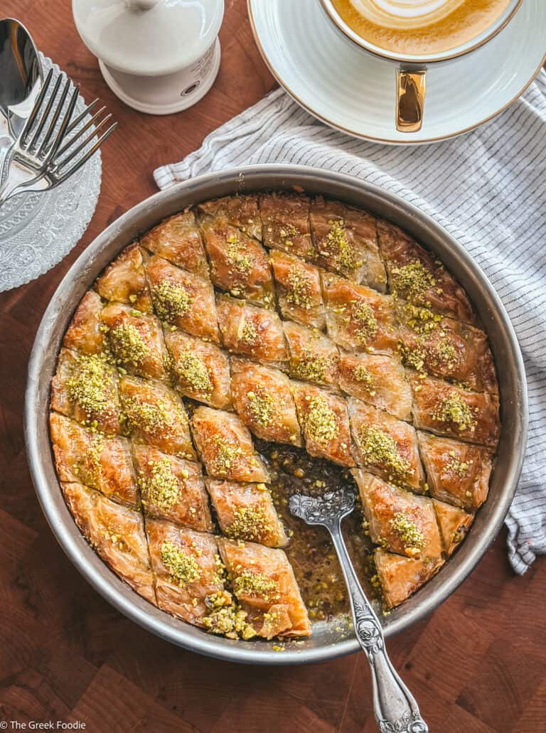 Overhead view of pistachio baklava in a round metal pan with a serving spoon, glass plates beside the baking pan and a cup with cappuccino coffee.