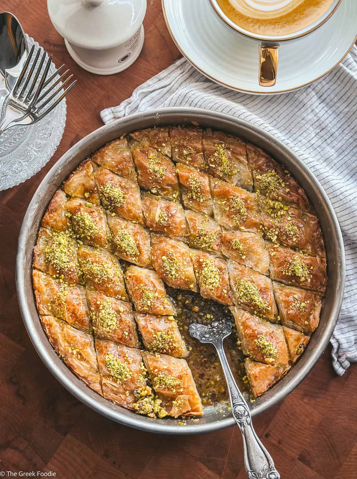 Overhead view of pistachio baklava in a round metal pan with a serving spoon, glass plates beside the baking pan and a cup with cappuccino coffee.