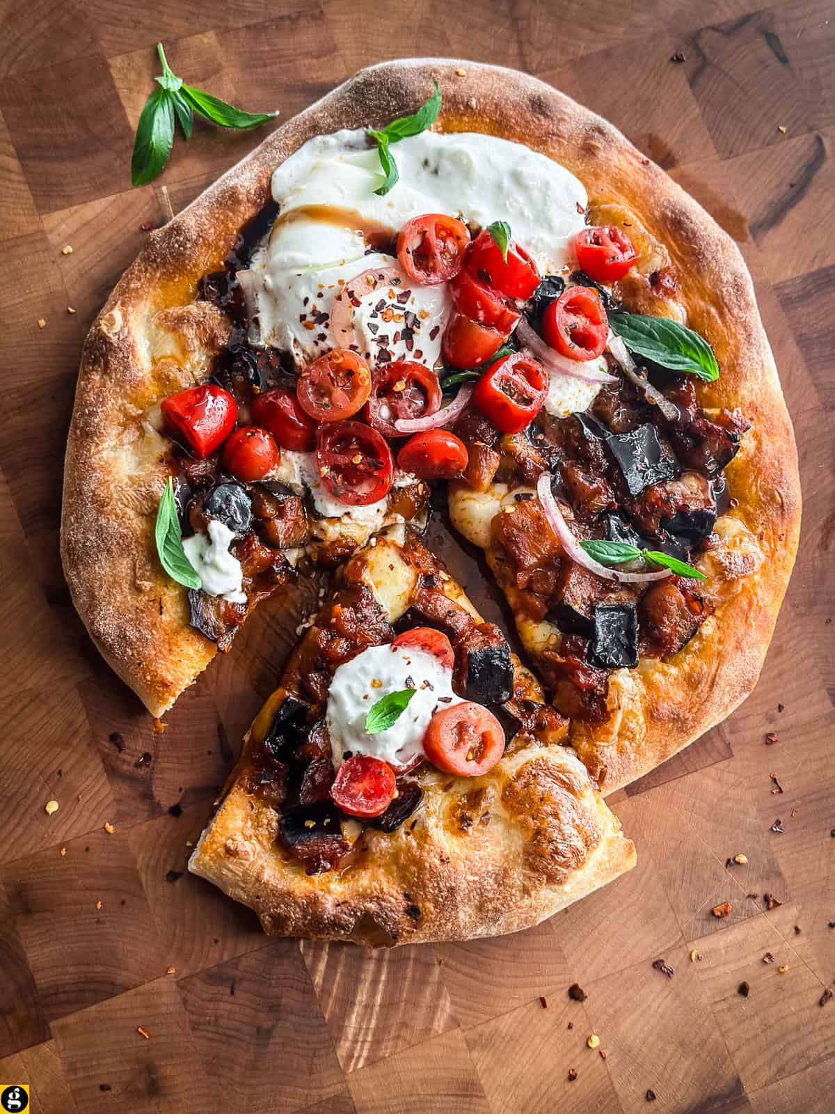 Sliced burrata eggplant pizza scattered with fresh basil and chili flakes on a butcher block.