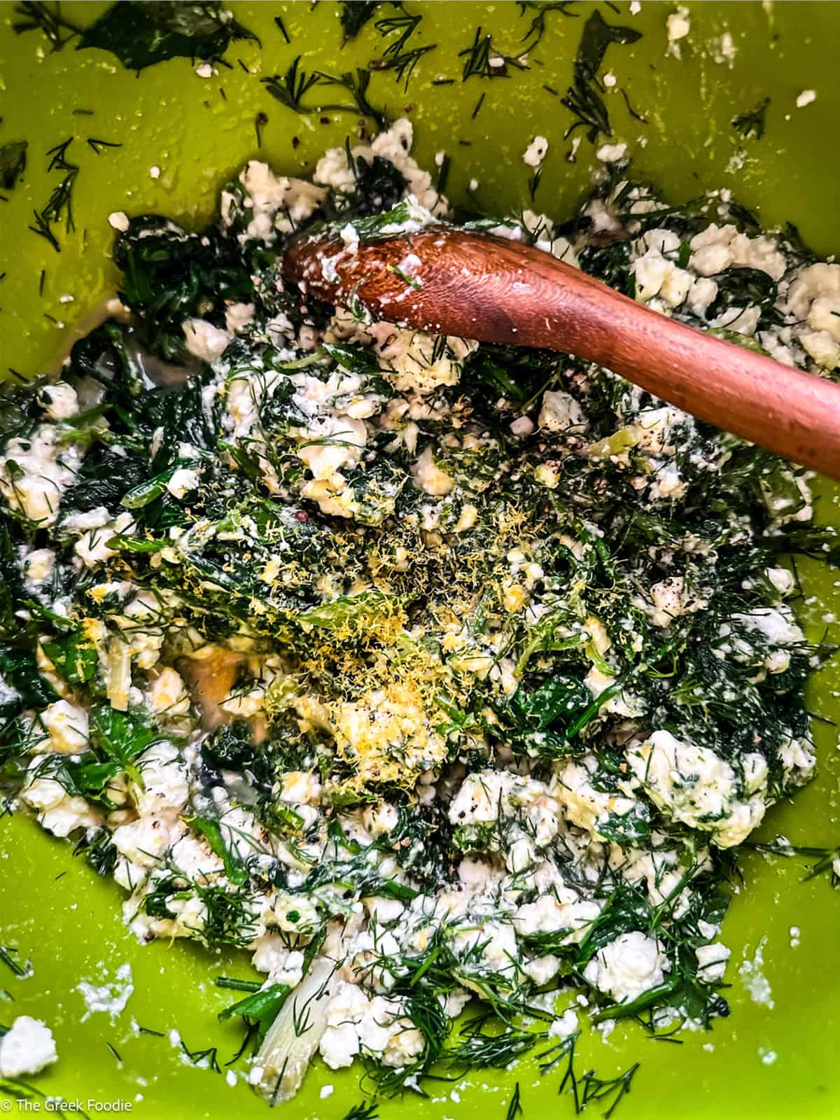 Sautéed spinach, crumbled feta, dill and seasoning mixed in a green bowl with a wooden spoon.