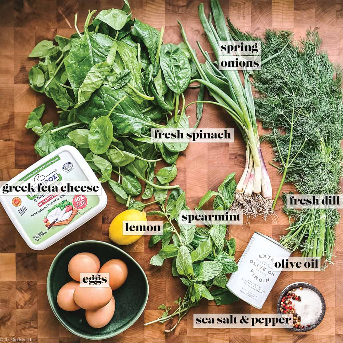 Ingredients for spanakopita on a wooden board: fresh spinach, spring onions, dill, spearmint, lemon, Greek feta, eggs, olive oil, sea salt and pepper.