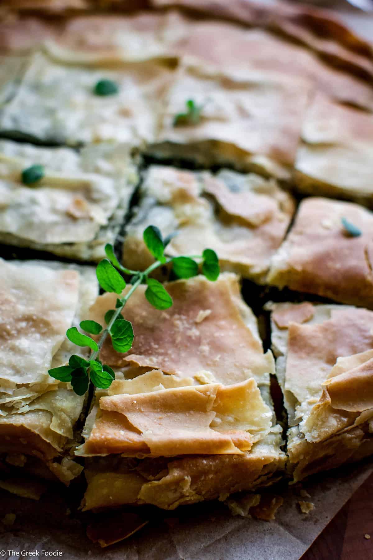 Close-up of spanakopita pieces with a sprig of fresh oregano across the golden phyllo.