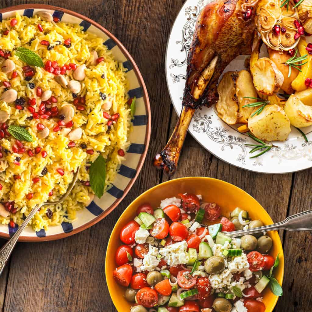 Greek side dishes on a wooden table: saffron rice pilaf with almonds and pomegranate, a Greek salad with feta and olives, and roasted potatoes.