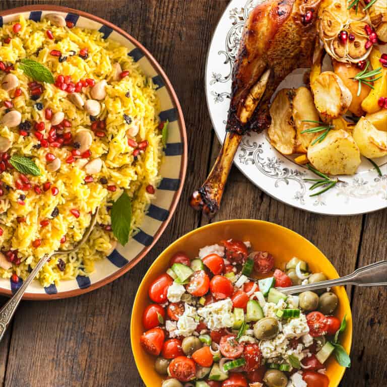 Greek side dishes on a wooden table: saffron rice pilaf with almonds and pomegranate, a Greek salad with feta and olives, and roasted potatoes.