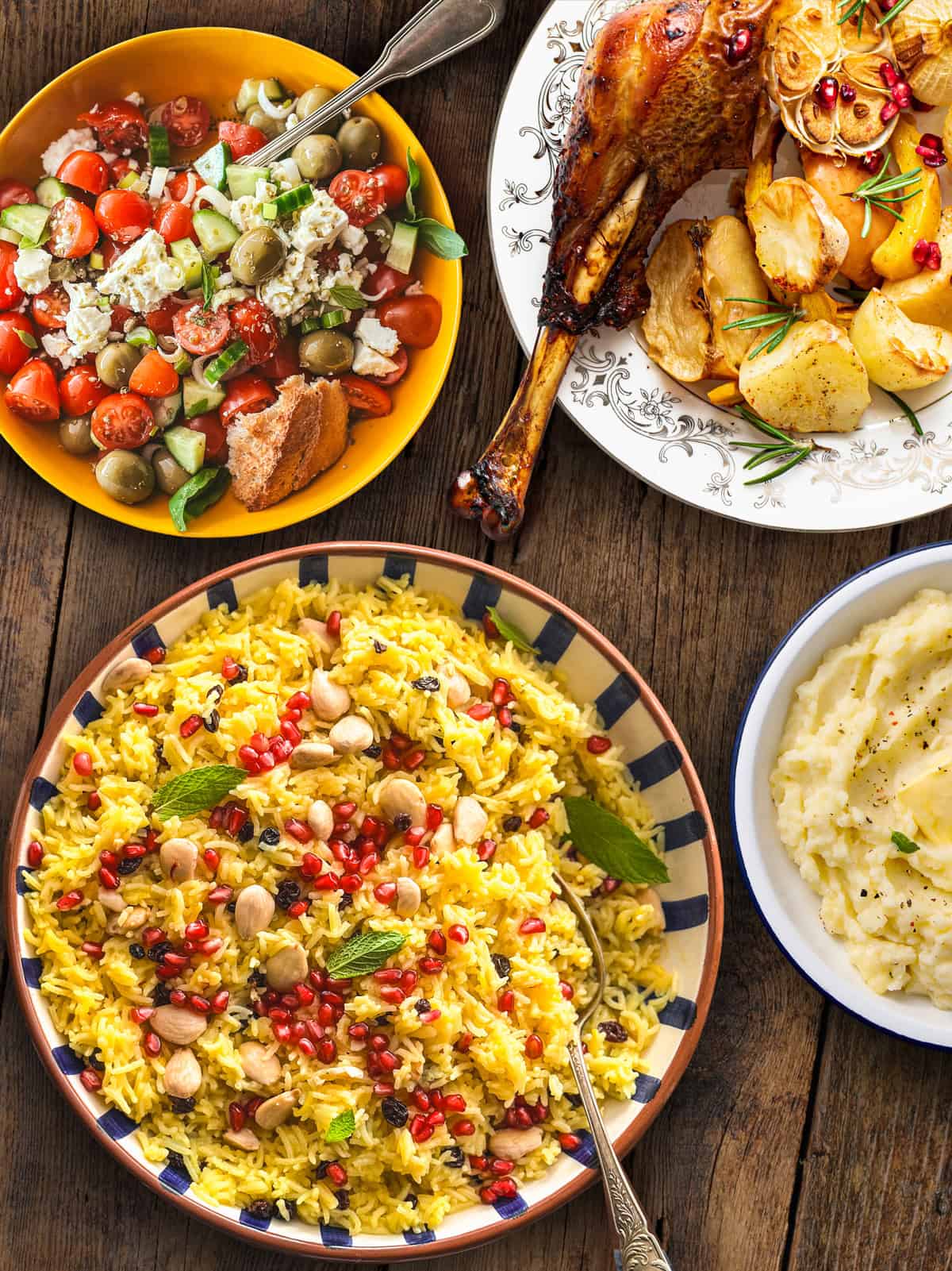 Greek side dishes on a wooden table: saffron rice pilaf with almonds and pomegranate, a Greek salad with feta and olives, olive oil mashed potatoes, and roast potatoes.